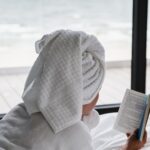 Lonely female with a white robe and a towel lying down in the bed and reading a book near the window