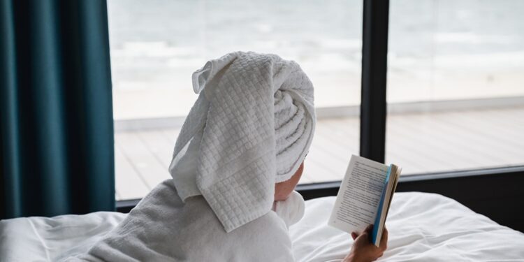 Lonely female with a white robe and a towel lying down in the bed and reading a book near the window