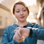 Young redhead woman wearing sportswear listening to music looking stopwatch at street