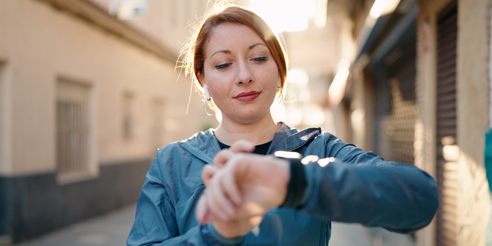 Young redhead woman wearing sportswear listening to music looking stopwatch at street